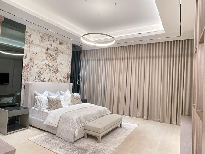 Modern bedroom with a neutral color palette, featuring a plush upholstered bed with decorative pillows, a marble accent wall, floor-to-ceiling curtains, and a stylish circular ceiling light.