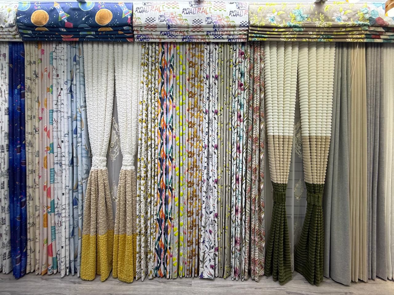 A photo of a rack displaying various curtain samples. The samples show a variety of colors, patterns, and textures, including solids, stripes, and floral prints.