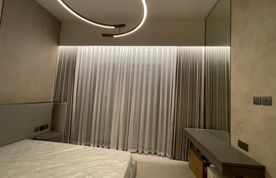 A photo of a modern bedroom with a large bed and a dresser. The room has a high ceiling with a circular light fixture and a large window with sheer curtains. The curtains let in natural light and create a soft, airy atmosphere in the room.